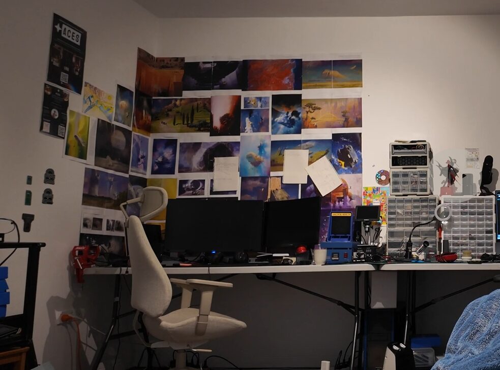 A desk with two monitors, parts drawers, and a wall of prints, with an ACES poster