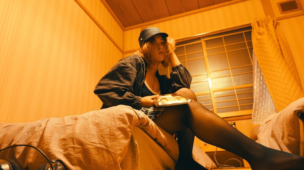 A warm wood room with a shoji screen, Freesia in a denim jacket and cap, takeaway box on her lap, headphones on the bed