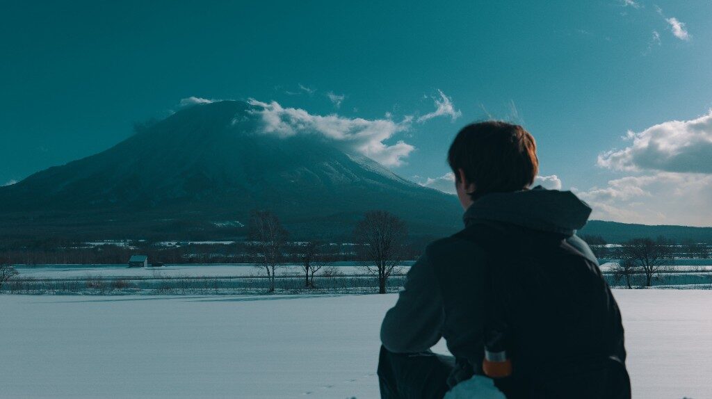 A snow field and a clouded mountain, seen from behind someone sitting in a dark jacket