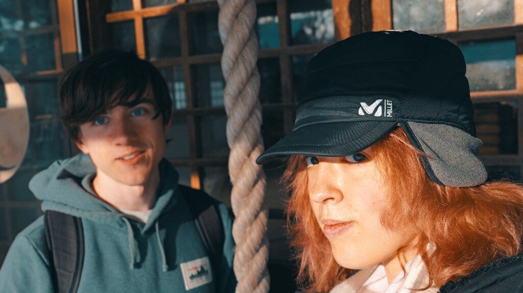 Freesia in a black cap beside a thick rope and a wooden window, with a friend in a green hoodie
