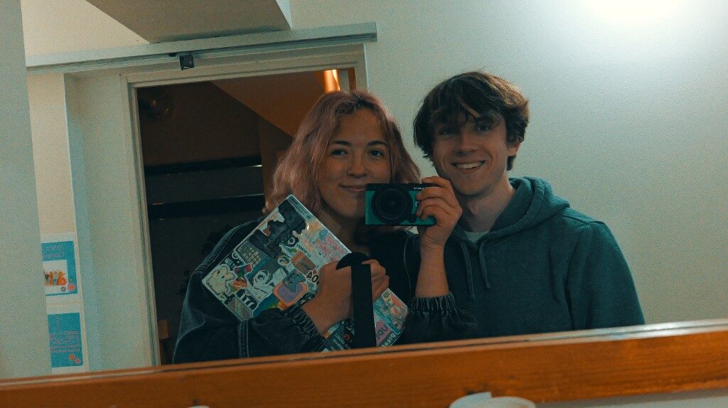 A mirror in a stairwell: Freesia with pink hair holding a stickered case, a friend holding a small camera