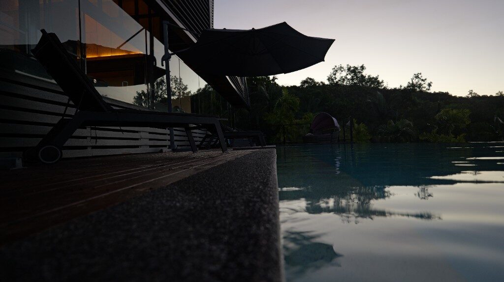 A pool at dusk beside a dark glass house, lounge chairs and a closed umbrella on the deck