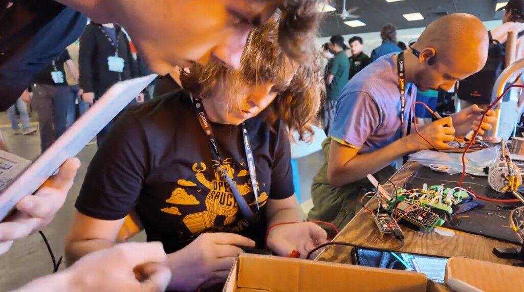 At a crowded workbench, wiring a board while people solder beside her