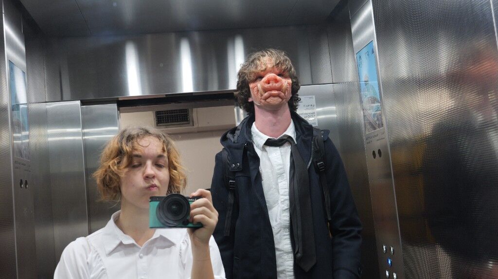 In a steel lift, Freesia with a small camera beside someone in a pig mask, shirt, and tie