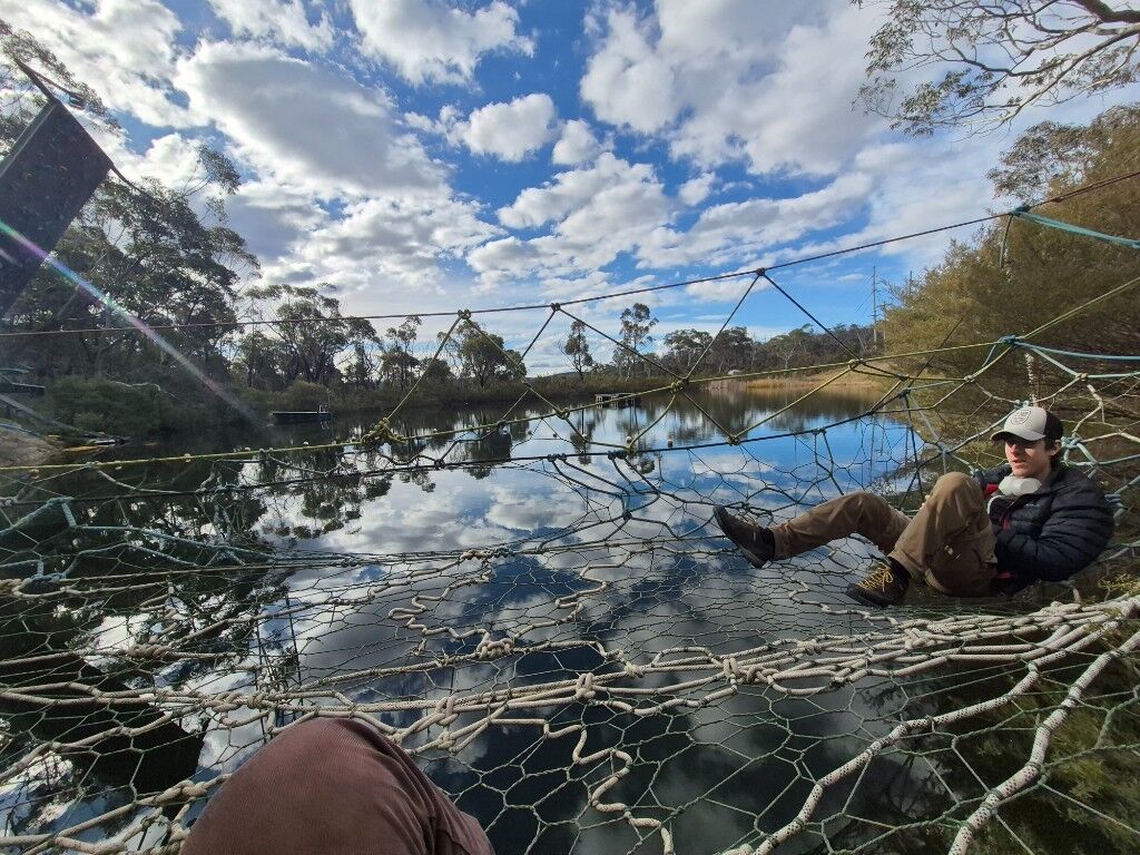 A rope net stretched over still water, someone sitting on it under a bright sky