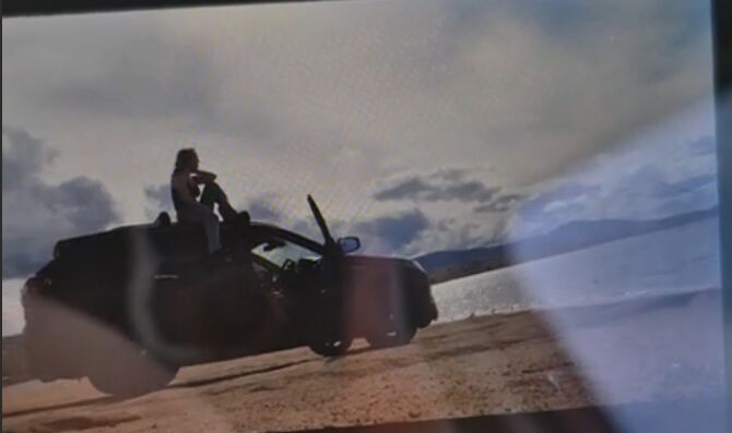 A photo of a screen: someone perched on a car by the water, doors open