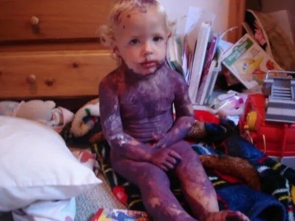 A small child covered in purple paint, sitting on a blanket by a dresser